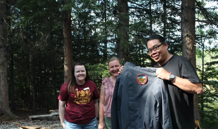 Norm Wong holds up a black Camp Chief Hector YMCA jacket, smiling alongside two other people.
