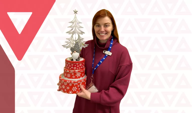 Lisa Rushton poses with the YMCA Calgary holiday gnome in front of a graphic background of YMCA triangles