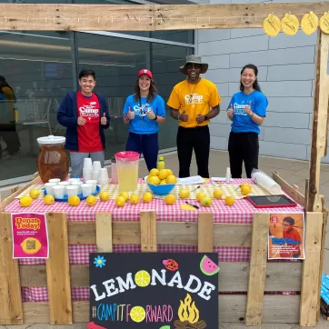 A group of 4 people stand in front of a wooden lemonade stand, smiling, in colourful t-shirts, to support the Camp It Forward campaign at YMCA Calgary.