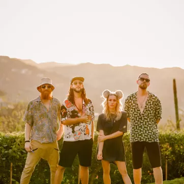 Walk Off the Eart, a band, stands in front of a mountain backdrop at golden hour, with 3 of the 4 members wearing sunglasses