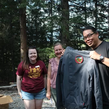 Norm Wong holds up a black Camp Chief Hector YMCA jacket, smiling alongside two other people.