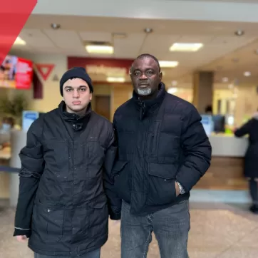 Two gentlemen stand in front of the Member Services desk at Saddletowne YMCA, both wearing black jackets and looking at the camera.
