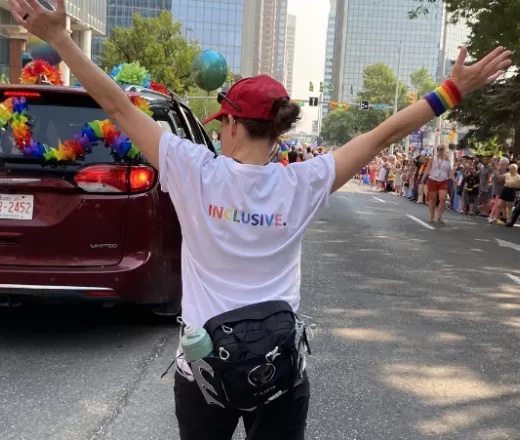 Individual with arms raised in the shape of a y with a white t shirt on that says inclusive on the back